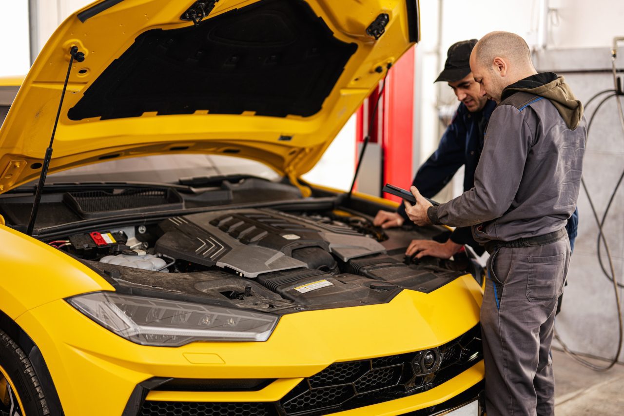 two mechanics stand with tbalet against open hood of yellow sport car suv .jpg